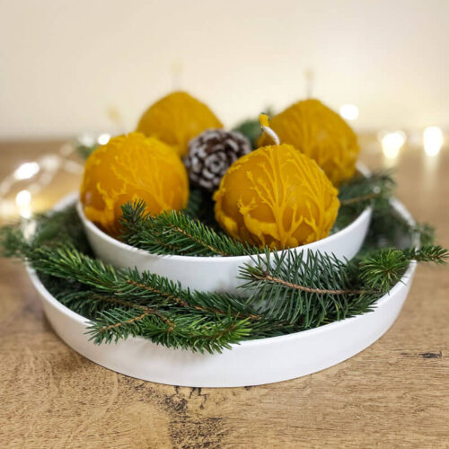 Gömb formájú méhviasz adventi gyertyák fehér tálban, karácsonyi fényekkel és természetes fenyőág díszítéssel.